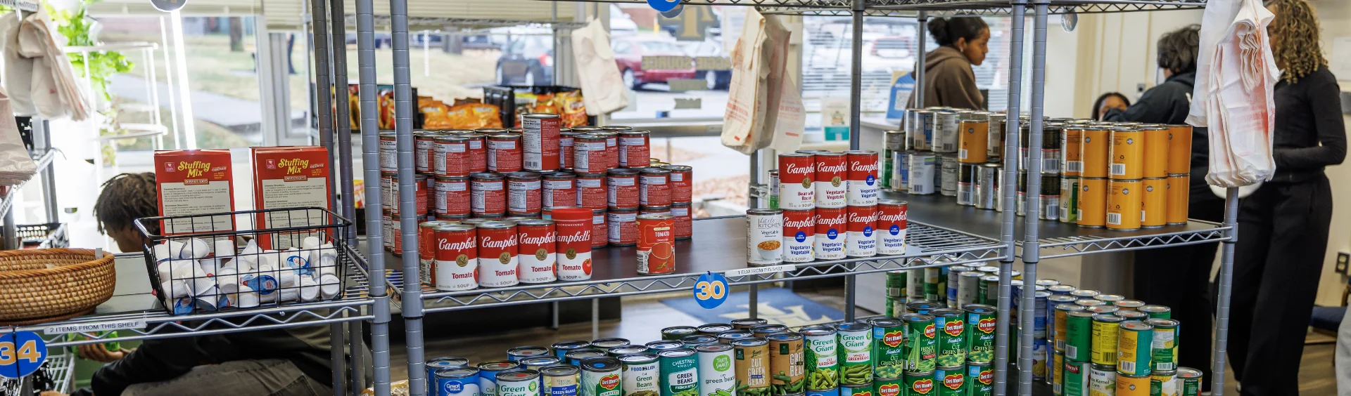 Metal wire shelving units in a community food pantry stocked with rows of Campbell's tomato soup, Green Giant vegetables, stuffing mix, and coffee pods, with people browsing the selection in the background