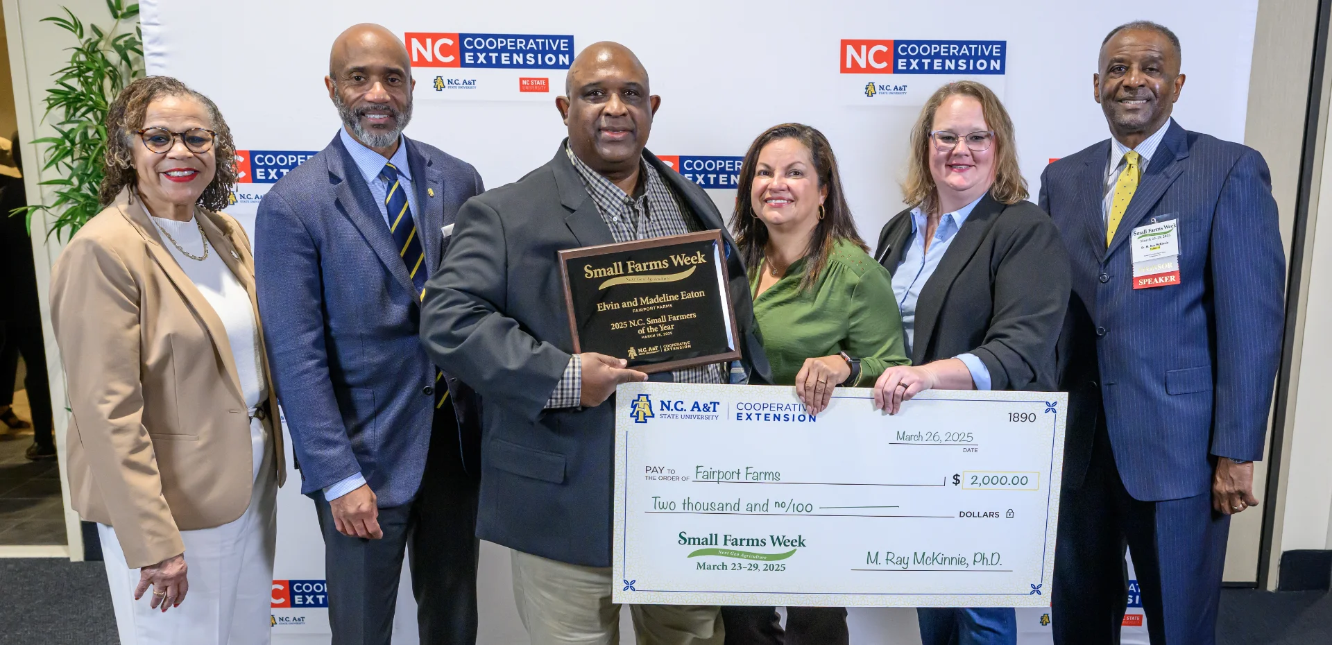 Seven people stand in front of an NC Cooperative Extension backdrop. The man in the center holds a plaque recognizing Elvin and Madeline Eaton of Fairport Farms as the 2025 N.C. Small Farmers of the Year. A large ceremonial check for $2,000 from N.C. A&T State University is also displayed.