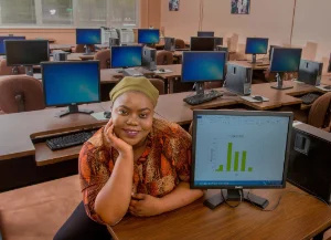 A smiling woman poses with her chin resting on her hand in front of a desktop computer displaying a bar chart in a computer lab filled with unoccupied workstations.
