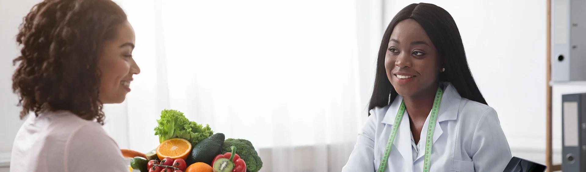 A nutritionist wearing a white coat and measuring tape around her neck talks with a woman holding a basket of fresh fruits and vegetables.