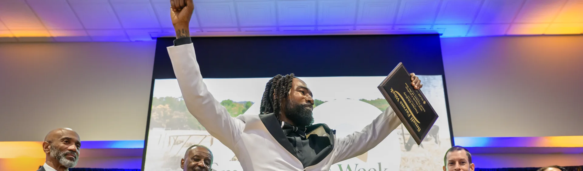 A man with dreadlocks wearing a white tuxedo jacket and black bowtie raises one arm in triumph while holding a 2026 Small Farmer of the Year award plaque in his other hand. He is smiling on a stage in front of a large screen during Small Farms Week.