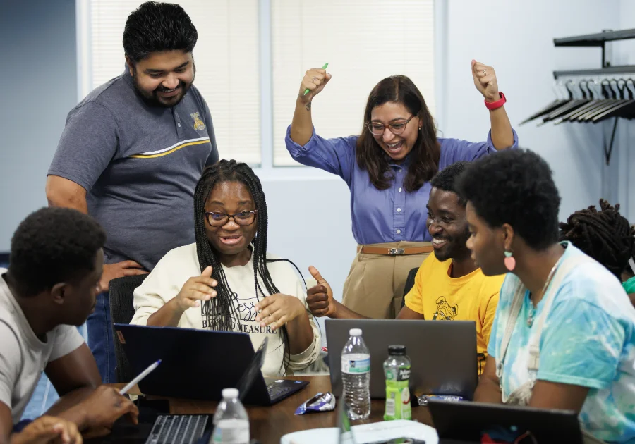 A group of students and instructors sit around a table with laptops, smiling and celebrating a success. One student gestures excitedly while a woman standing behind them raises her arms in triumph.